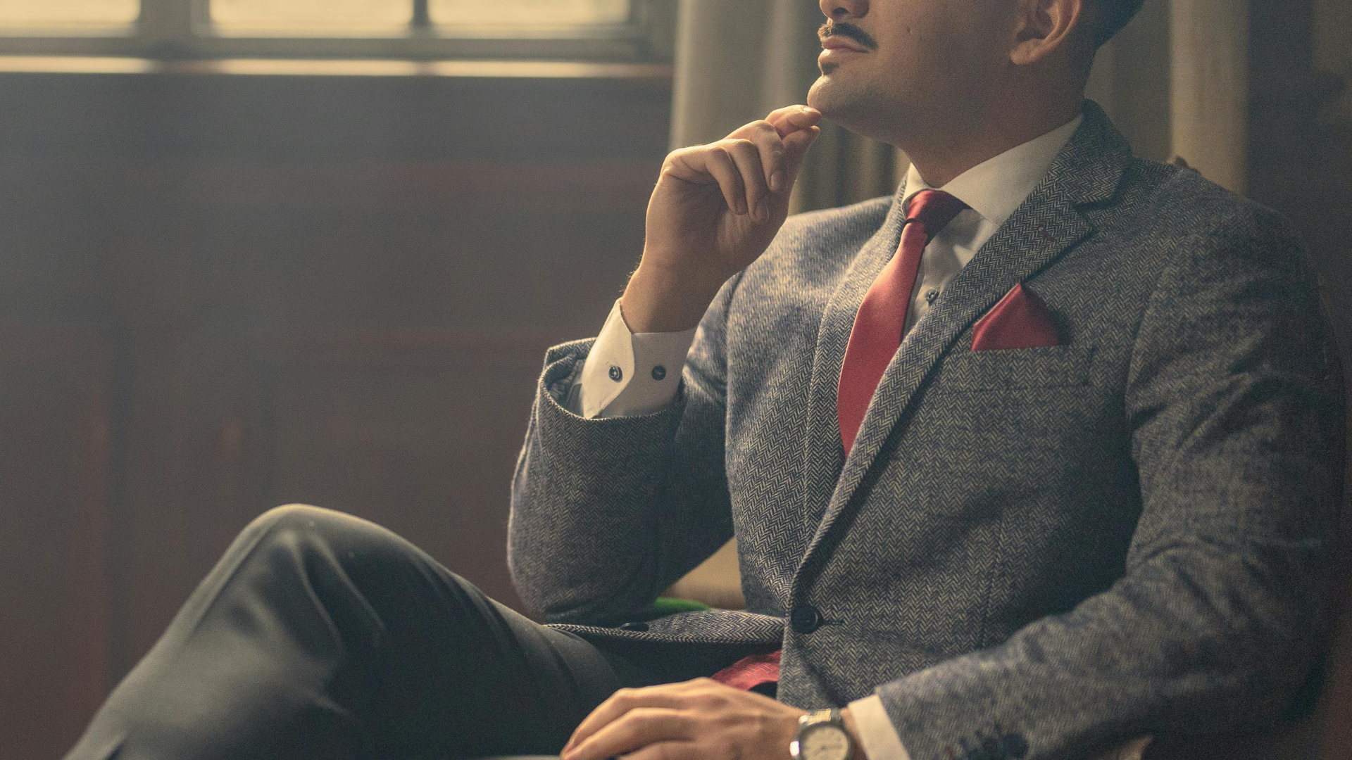 グレーのツイードジャケットと赤いネクタイを着用し、髭をたくわえた男性